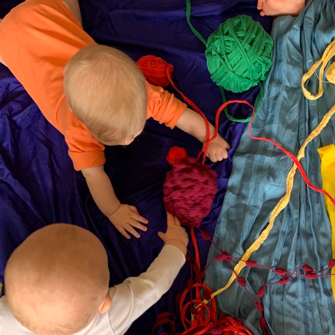 Babysar ligger på mage och leker med tyg och garner. 