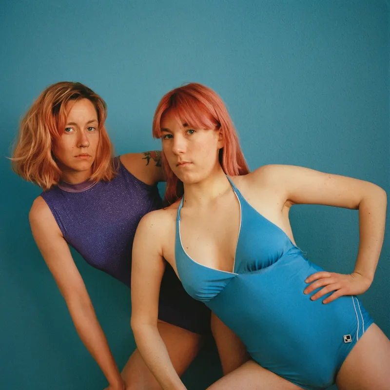 Två kvinnor poserar i baddräkt framför en blå bakgrund. 