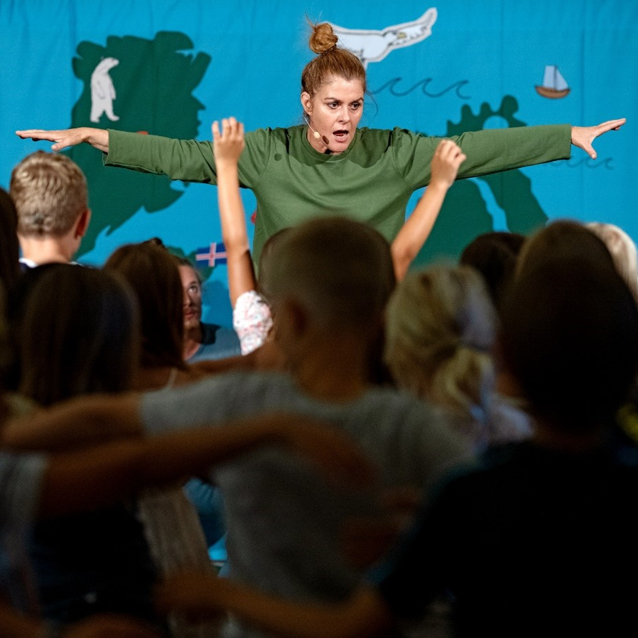 En kvinna i grön tröja står på en scen framför en publik av barn. 
